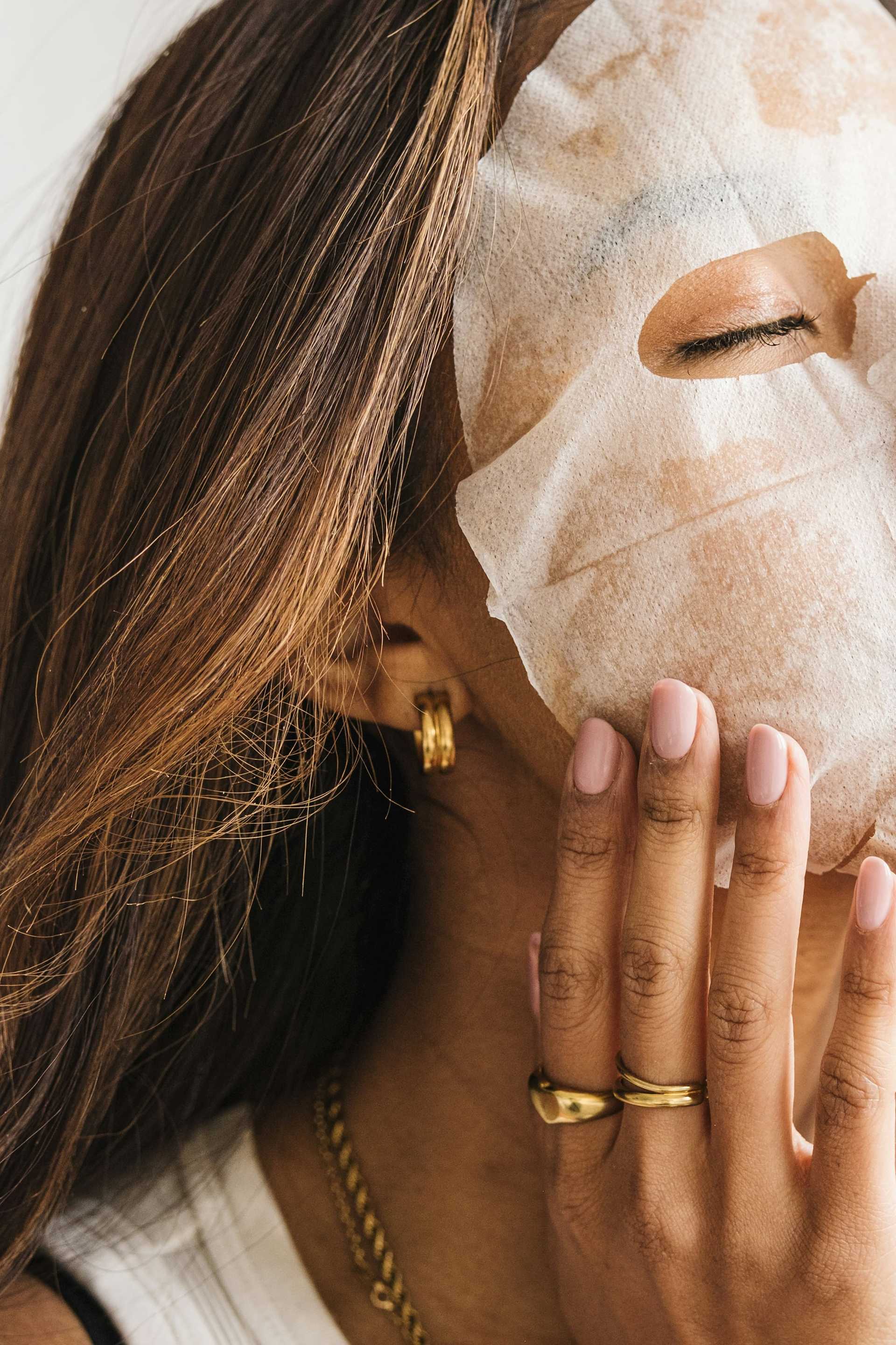 Woman with a sheet mask, touching face, wearing gold jewelry.